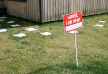 Czerwony szyld „House for rent” wbity na trawniku przed domem.
