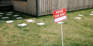 Czerwony szyld „House for rent” wbity na trawniku przed domem.