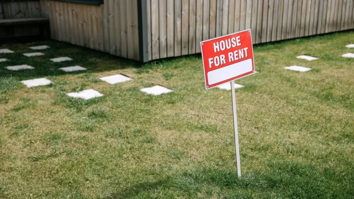 Czerwony szyld „House for rent” wbity na trawniku przed domem.